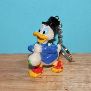May include: A keychain featuring a cartoon character duck wearing a black top hat and blue jacket, holding a money bag. The duck has a yellow beak and red and yellow shoes. The keychain is attached to a silver chain and ring.