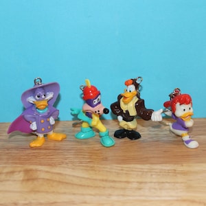 Könnte beinhalten: Vier bunte Schlüsselanhänger mit Zeichentrickfiguren. Die Figuren umfassen eine lila Ente mit Umhang, eine hundeartige Figur mit rotem Helm, eine Ente in einer braunen Jacke und eine Ente mit roter Schleife. Sie stehen auf einer Holzoberfläche.