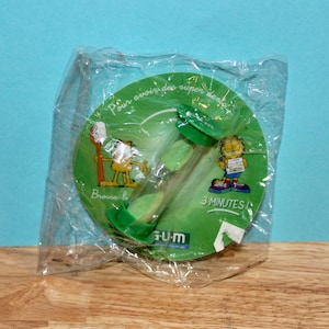 Può includere: Un timer a clessidra verde da 3 minuti con un design a cartoni animati di Garfield, confezionato in plastica trasparente. Il timer ha tappi terminali verdi ed è posto su una base circolare verde con testo e grafica di Garfield. La confezione include il marchio "GUM".