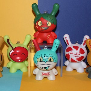 May include: Seven colorful, cartoon-like, plush toys with different designs. The toys are all about 4 inches tall and have a loop for hanging. The toys are on a yellow and blue surface.