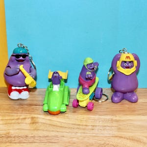 May include: Set of four purple cartoon character keychains. The characters are wearing different outfits and accessories, including a hat, sunglasses, a skateboard, and a race car.