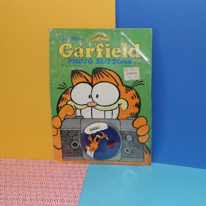 May include: A Garfield photo button featuring Garfield in a blue shirt with the text "Banzai!" on a blue background. The button is packaged in a plastic bag with a cardboard backing featuring Garfield and the text "Garfield Photo Buttons".