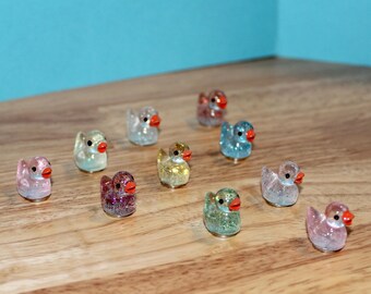 Puede incluir: Una colección de pequeñas figuras de patos brillantes en varios colores, incluyendo rosa, azul y dorado. Cada pato tiene un pico rojo y está dispuesto sobre una superficie de madera. Los patos son un artículo decorativo caprichoso.