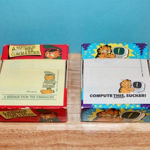 May include: Two rectangular paper note pads in cardboard boxes featuring Garfield. The left box is red with the text A MESSAGE FROM THE COMMANDER. The right box is blue with yellow accents and the text COMPUTE THIS, SUCKER! both include illustrations.