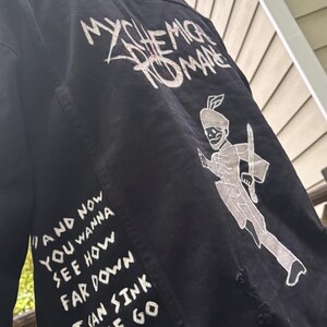 Op de afbeelding: Een zwart spijkerjack met witte zeefdruktekst en afbeeldingen op de achterkant. De tekst luidt My Chemical Romance en de songtekst And now you wanna see how far down I can sink let me go fuck. Een skeletachtige figuur is ook afgebeeld.