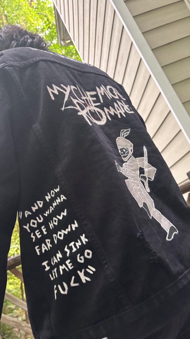 Op de afbeelding: Een zwart spijkerjack met witte zeefdruktekst en afbeeldingen op de achterkant. De tekst luidt My Chemical Romance en de songtekst And now you wanna see how far down I can sink let me go fuck. Een skeletachtige figuur is ook afgebeeld.