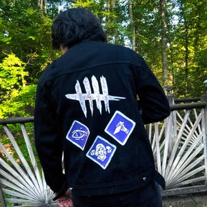 May include: A black denim jacket featuring a white hand-painted logo of five vertical slashes at the top. Below the logo are three purple diamond-shaped patches with white illustrations of an eye, a hand holding a knife, and a person with skulls.