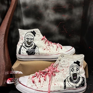 May include: White high-top trainers with a horror clown design. The trainers feature a black and white illustration of a clown face on the sides, with red splatter details and matching laces. The shoes are displayed on a brown surface.