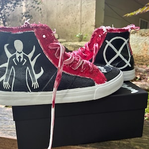 May include: High-top trainers featuring a black canvas base with a white graphic of a tall figure with tentacles on the side. The shoes have red frayed fabric accents along the eyelets, white laces, and a white rubber sole. The heel displays a white circle with an X.
