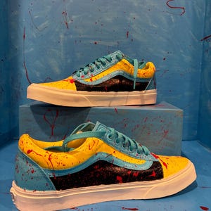 Può includere: Un paio di scarpe da ginnastica dipinte su misura. Le scarpe presentano una combinazione di colori giallo e blu con dettagli neri e spruzzi rossi. I lacci sono turchese e le suole bianche. Sono esposte su una superficie blu con spruzzi di vernice rossa.