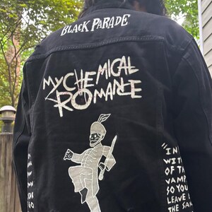 Op de afbeelding: Een zwart spijkerjack met witte zeefdrukafbeeldingen op de achterkant. De tekst luidt "BLACK PARADE" en "MY CHEMICAL ROMANCE" boven een afbeelding van een skelet in een marsorkest. Songteksten zijn gedrukt op de zijkanten van de achterkant.