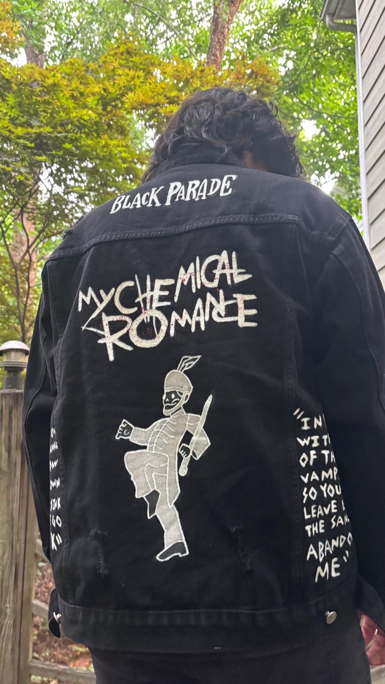 Op de afbeelding: Een zwart spijkerjack met witte zeefdrukafbeeldingen op de achterkant. De tekst luidt "BLACK PARADE" en "MY CHEMICAL ROMANCE" boven een afbeelding van een skelet in een marsorkest. Songteksten zijn gedrukt op de zijkanten van de achterkant.