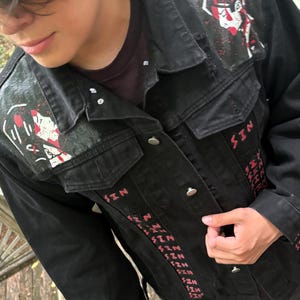 Op de afbeelding: Een zwart spijkerjack met handgeschilderde witte en rode grafische ontwerpen op de schouders en de herhaalde rode tekst "SIN" verticaal gedrukt langs de sluiting aan de voorzijde. Het jack heeft een klassieke kraag en metalen knopen als sluiting.