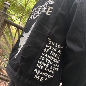 Op de afbeelding: Een zwart spijkerjack met handgeschilderde witte tekst die "IN LOVE WITH ALL OF THESE VAMPIRES SO YOU CAN LEAVE LIKE THE SAME ABANDON ME" en "MY CHEMICAL ROMANCE" leest, samen met een witte grafische illustratie van een skeletfiguur.