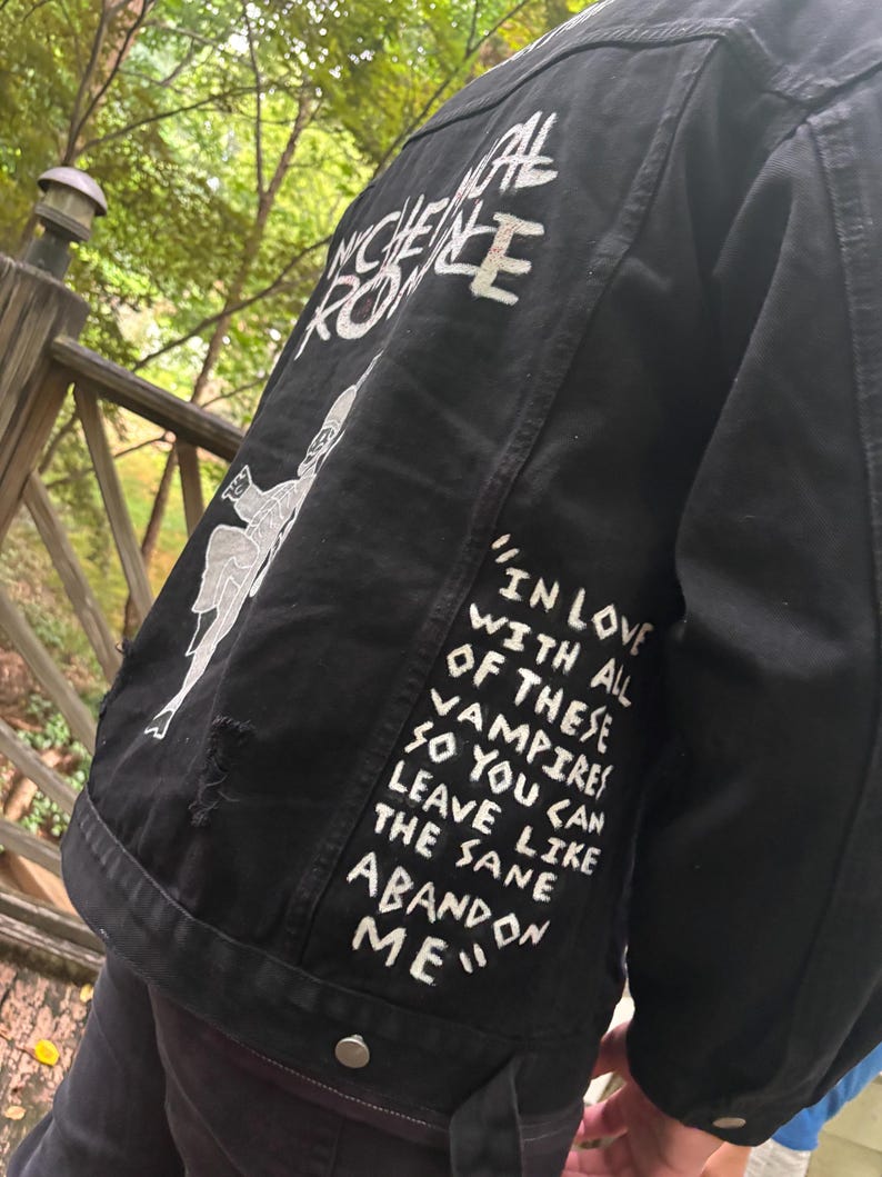 Op de afbeelding: Een zwart spijkerjack met handgeschilderde witte tekst die "IN LOVE WITH ALL OF THESE VAMPIRES SO YOU CAN LEAVE LIKE THE SAME ABANDON ME" en "MY CHEMICAL ROMANCE" leest, samen met een witte grafische illustratie van een skeletfiguur.