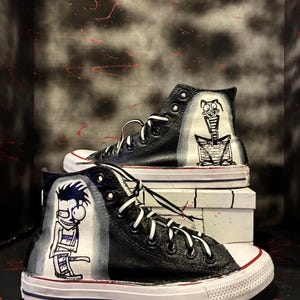 Puede incluir: Zapatillas altas negras con cordones blancos y suela blanca. Las zapatillas presentan personajes de dibujos animados pintados a mano en blanco y negro en los laterales. Los zapatos se exhiben sobre una superficie blanca sobre un fondo oscuro.