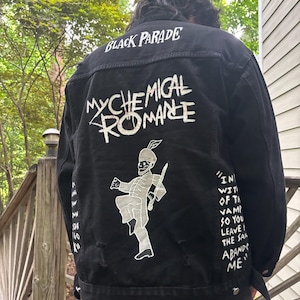 Op de afbeelding: Een zwart spijkerjack met witte zeefdrukafbeeldingen op de achterkant. De tekst luidt "BLACK PARADE" en "MY CHEMICAL ROMANCE" boven een figuur van een marsmuzikant. De tekst aan de rechterkant luidt "IN WITH OF THE VAMP SO YOU LEAVE THE SAME ABAND ME".