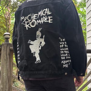 Op de afbeelding: Een zwart spijkerjack met witte zeefdrukafbeeldingen op de achterkant. De tekst luidt BLACK PARADE en MY CHEMICAL ROMANCE, vergezeld door een figuur van een marsmuzikant en songteksten met de woorden IN WITH THE VAMPIRES SO YOU LEAVE.