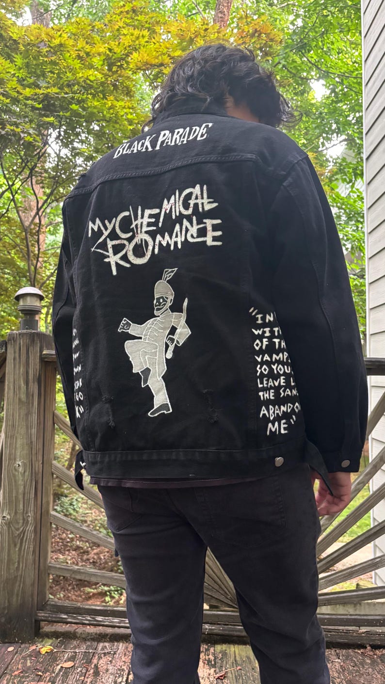 Op de afbeelding: Een zwart spijkerjack met witte zeefdrukafbeeldingen op de achterkant. De tekst luidt BLACK PARADE en MY CHEMICAL ROMANCE, vergezeld door een figuur van een marsmuzikant en songteksten met de woorden IN WITH THE VAMPIRES SO YOU LEAVE.
