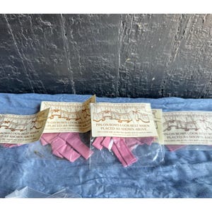Op de afbeelding: Meerdere doorzichtige plastic verpakkingen met roze stoffen strikken en veiligheidsspelden. Elke verpakking heeft een wit papieren label met een bruine lijntekening van een gordijn en de tekst: PIN-ON BOWS LOOK BEST WHEN PLACED AS SHOWN ABOVE.