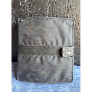 May include: A black Mead Five Star zippered binder featuring a horizontal fabric strap across the center with a rectangular closure tab. The tab displays the text Five Star with five stars underneath. The binder has a small Mead brand tag.