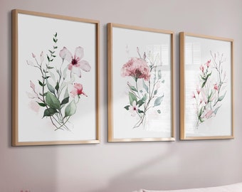 May include: Three framed wall art prints featuring watercolor floral illustrations in shades of pink and green. Each print is encased in a light wood frame and displayed in a row on a wall above a light pink sofa with matching cushions.