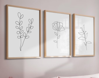 May include: Three framed line drawings of floral designs in black on a white background. The frames are light wood and the drawings are hung on a light pink wall above a light pink sofa with three pillows.