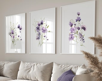 May include: A set of three framed wall art prints featuring watercolour illustrations of purple wildflowers and green stems. The prints are displayed in thin white frames hanging in a row above a large beige sofa with purple throw pillows.