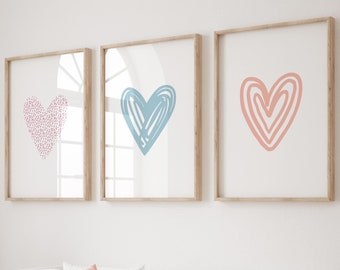 Puede incluir: Tres impresiones artísticas enmarcadas con diseños de corazón en rosa, azul y coral cuelgan sobre una cama blanca con una manta y almohadas color coral. Un caballito de madera y una cesta de mimbre están en primer plano. La habitación tiene una paleta de colores neutros.