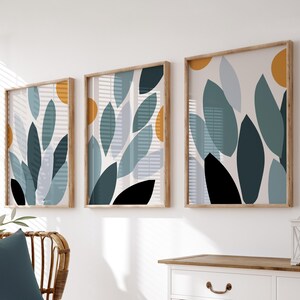 May include: Three framed abstract art prints with botanical leaf designs in teal, black, and light blue, accented with orange circles. The prints are displayed on a white wall above a white cabinet and a wicker chair with a blue cushion and white blanket.