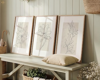 Op de afbeelding: Een lichtgroene houten bank bevat drie ingelijste prints met zwarte lijntekeningen van bloemen op een beige achtergrond. Een beige kussen ligt op de bank, met een gevlochten mand met een doek en een plant eronder. Een bloemenkrans hangt erboven.