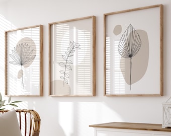 May include: Three framed illustrations of stylized plants with brown and beige accents on a white wall. A wicker chair with a beige cushion and a white throw blanket sits in front of the artwork. A white cabinet with a wooden top and two drawers is to the right of the chair. A wicker basket sits on the floor in front of the cabinet.
