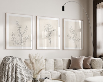 May include: Three framed wall art prints featuring black line drawings of botanical stems and flowers on a light beige background. The prints are displayed in a row above a light-colored sofa in a room with a window and a floor lamp.