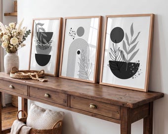 May include: Three framed wall art prints featuring black and gray abstract geometric shapes, lines, and botanical illustrations. The prints are displayed in light wood frames on a wooden console table next to a vase of dried flowers.