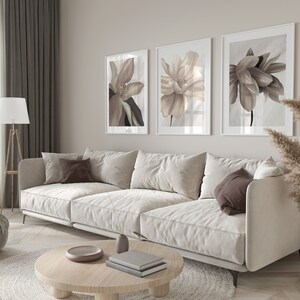 May include: A white sofa with brown throw pillows sits in a living room with three framed floral prints on the wall. A round wooden coffee table with books and a decorative bowl sits in front of the sofa. A beige rug is under the coffee table.