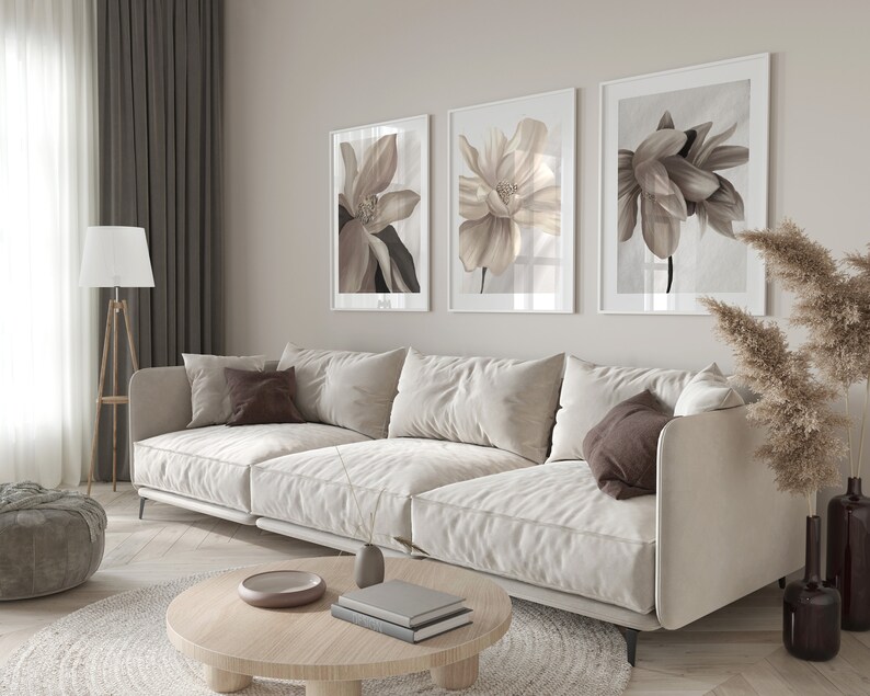 May include: A white sofa with brown throw pillows sits in a living room with three framed floral prints on the wall. A round wooden coffee table with books and a decorative bowl sits in front of the sofa. A beige rug is under the coffee table.