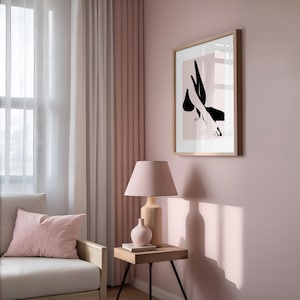 May include: A framed wall art print featuring a black abstract silhouette of legs and feet against a pale pink background. The artwork is displayed in a light wood frame, hanging on a solid light pink wall above a side table with a lamp.