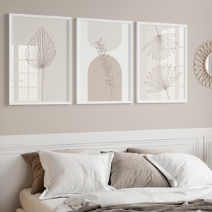 Puede incluir: Tres cuadros rectangulares en marcos blancos con dibujos de l&iacute;neas de hojas bot&aacute;nicas y formas geom&eacute;tricas en tonos beige y blanco. Los cuadros est&aacute;n dispuestos horizontalmente en una pared sobre una cama con almohadas blancas y marrones.