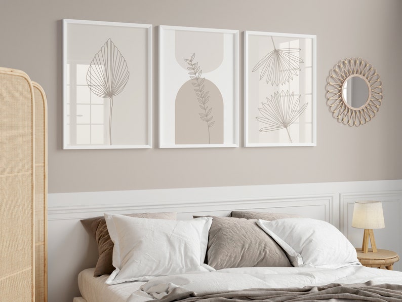 Puede incluir: Tres cuadros rectangulares en marcos blancos con dibujos de l&iacute;neas de hojas bot&aacute;nicas y formas geom&eacute;tricas en tonos beige y blanco. Los cuadros est&aacute;n dispuestos horizontalmente en una pared sobre una cama con almohadas blancas y marrones.