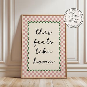May include: A framed art print with a pink and white checkered pattern. The central cream-colored rectangle displays the words "this feels like home" in black script. A green wavy border frames the text. A circular graphic in the upper right corner says "Choose your own colour".