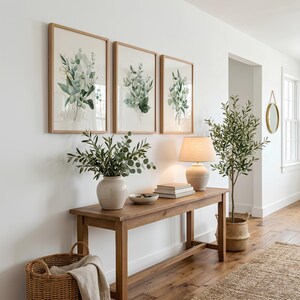 May include: Three framed botanical prints featuring green eucalyptus leaves in light wood frames, displayed above a wooden console table with a ceramic vase, a lamp, a stack of books, and a small bowl, alongside a potted olive tree.