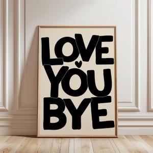 Könnte beinhalten: Ein gerahmter Kunstdruck mit den Worten "LOVE YOU BYE" in fetten, schwarzen Buchstaben auf cremefarbenem Hintergrund. Ein kleines schwarzes Herz befindet sich zwischen "YOU" und "BYE". Der Druck ist in einem hellbraunen Holzrahmen.