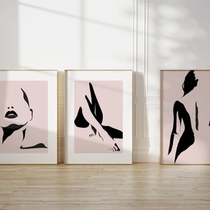 May include: Three framed wall art prints featuring black ink-style silhouettes on a pale pink background. The first print shows a face, the second shows legs, and the third shows a back. Each piece is enclosed in a thin, light-colored wooden frame.