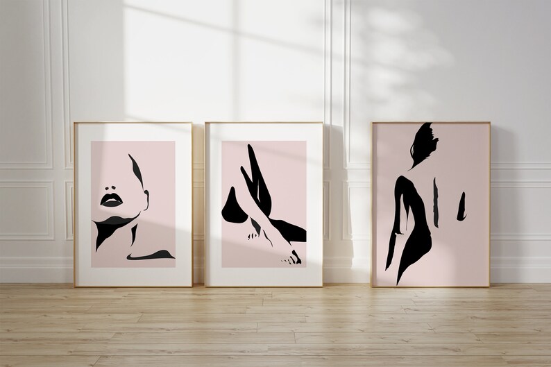 May include: Three framed wall art prints featuring black ink-style silhouettes on a pale pink background. The first print shows a face, the second shows legs, and the third shows a back. Each piece is enclosed in a thin, light-colored wooden frame.