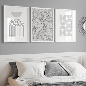 May include: Three black and white line art prints in white frames on a gray wall above a bed with white bedding and gray pillows. The prints feature abstract geometric shapes, a floral design with faces, and an arch.