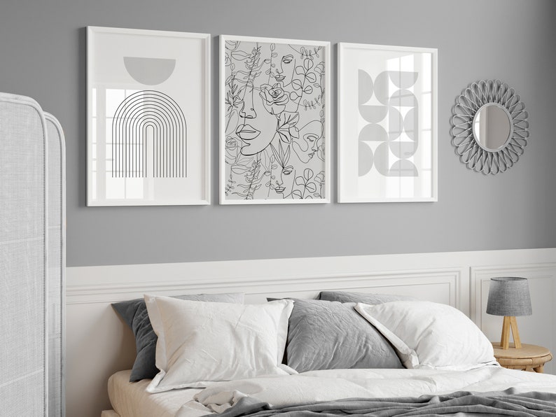 May include: Three black and white line art prints in white frames on a gray wall above a bed with white bedding and gray pillows. The prints feature abstract geometric shapes, a floral design with faces, and an arch.