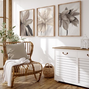May include: A white cabinet with two drawers and two doors with a wood top. The cabinet is decorated with a brown, wooden handle. The cabinet is in front of a white wall with three framed prints of beige flowers. A wicker chair with a beige cushion and a white throw blanket is in front of the cabinet. A wicker basket is on the floor next to the chair.
