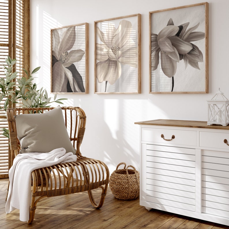 May include: A white cabinet with two drawers and two doors with a wood top. The cabinet is decorated with a brown, wooden handle. The cabinet is in front of a white wall with three framed prints of beige flowers. A wicker chair with a beige cushion and a white throw blanket is in front of the cabinet. A wicker basket is on the floor next to the chair.