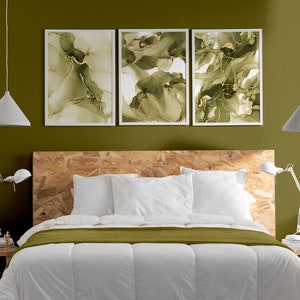 May include: A white bed with a green duvet cover and two white pillows. The headboard is made of wood and has a natural finish. There are three framed abstract paintings above the headboard, each with a green and gold design. Two white pendant lights hang from the ceiling.