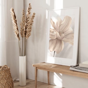 May include: A beige and white floral print in a white frame hangs on a white wall above a light wood bench. A vase with dried pampas grass sits on the floor next to the bench.